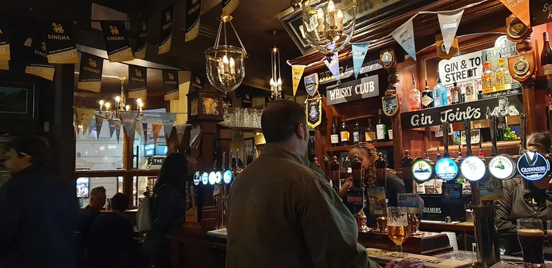 A small, warmly lit London pub interior with hanging lamps, bar taps and a customer at the counter