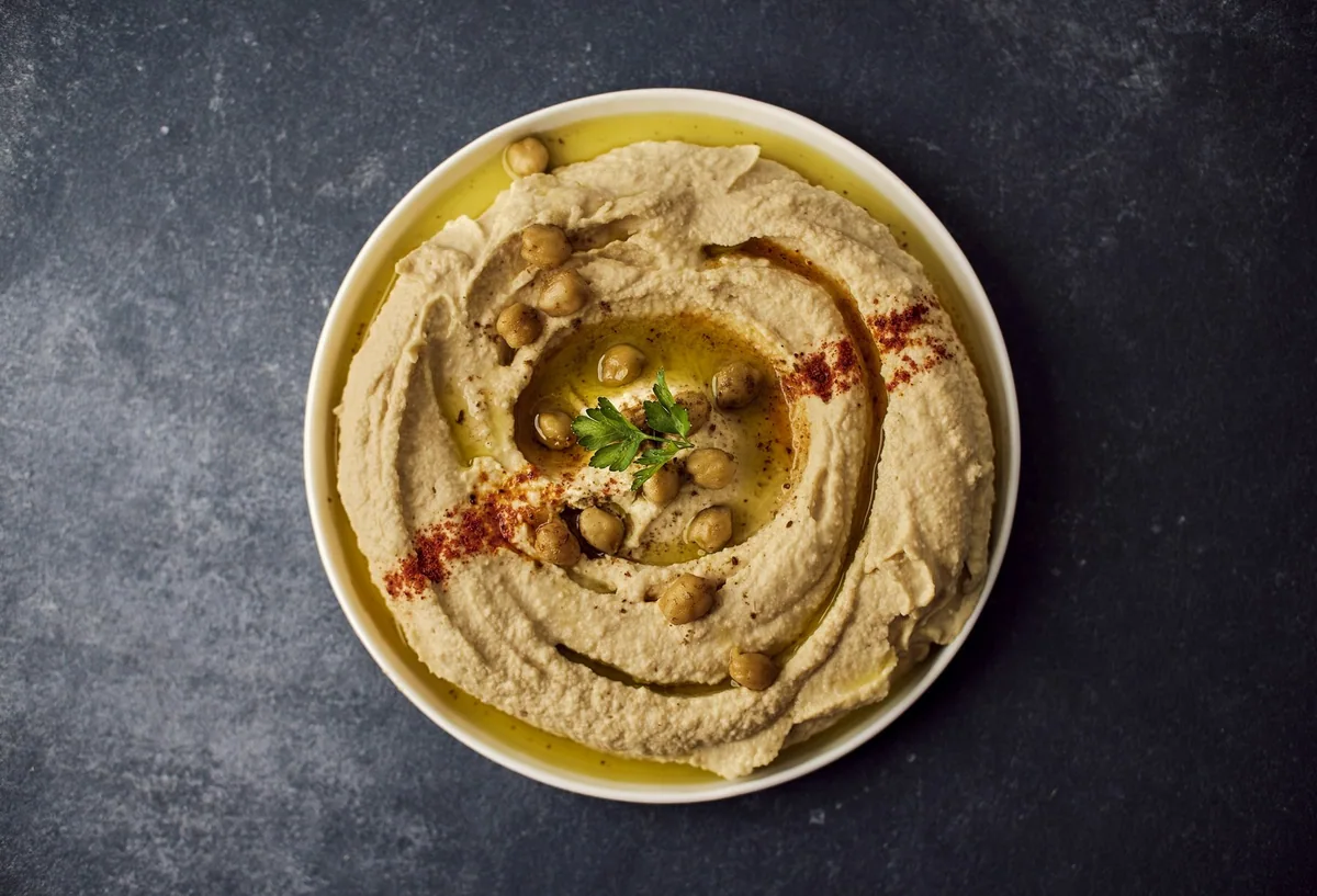 A bowl of hummus swirled with olive oil and paprika, topped with whole chickpeas and a sprig of parsley, seen from above