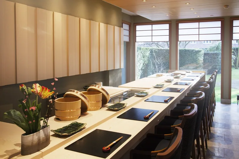 A long polished wooden sushi counter with stools drawn up to it and the chef's station behind