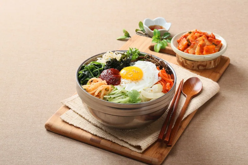 A bowl of bibimbap with a fried egg and cut vegetables over rice, served with a dish of radish kimchi