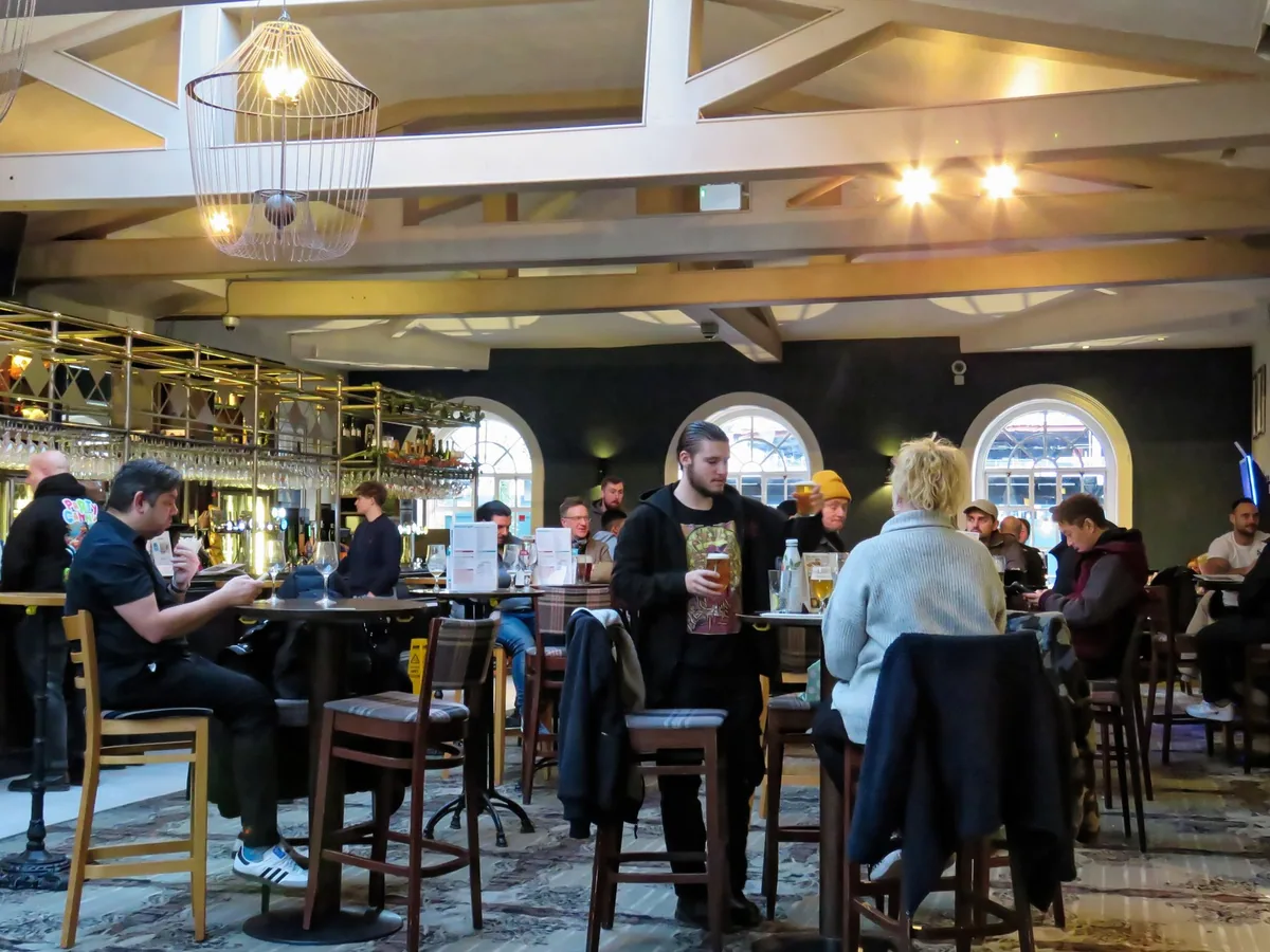 Drinkers at high tables in a busy London pub, one carrying two pints back from the bar