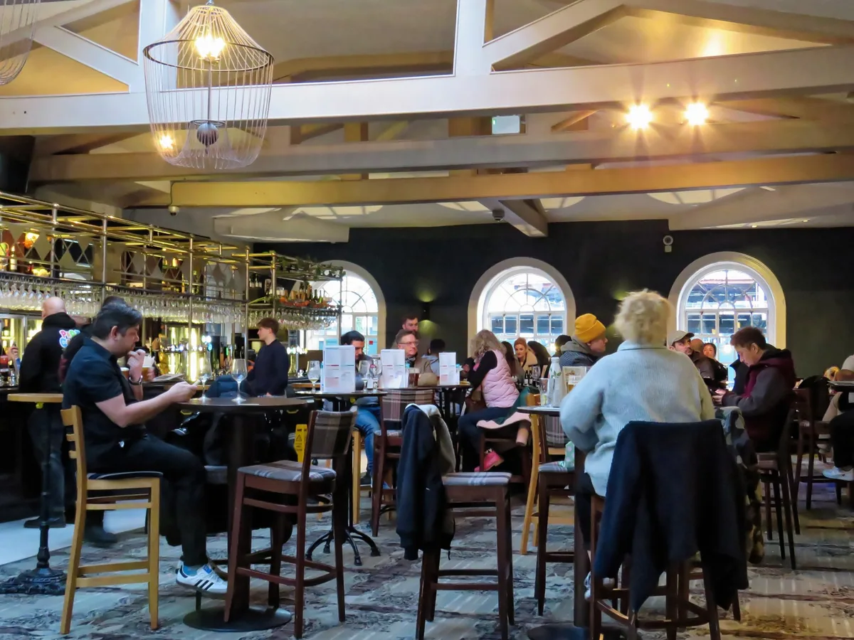 A busy London pub at West India Quay, with drinkers at high tables under tall arched windows