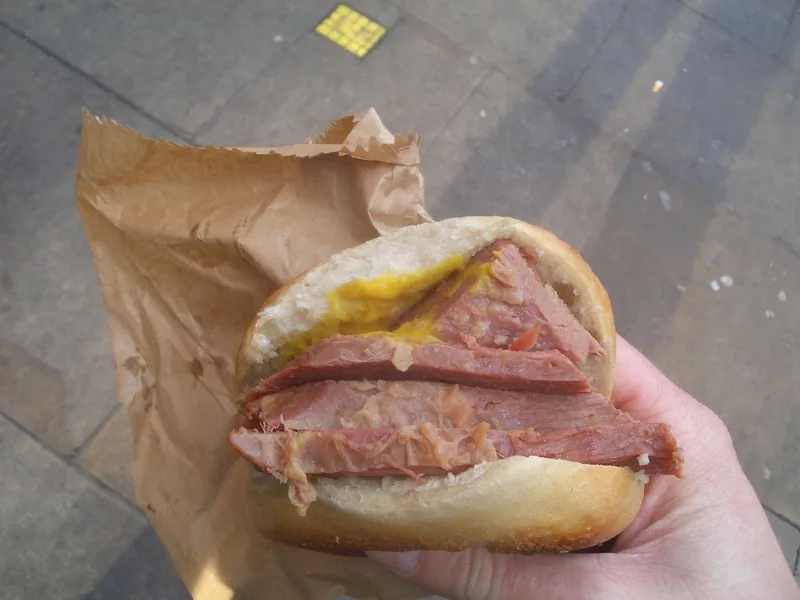 A salt beef beigel held in its paper wrapper, thick pink slices with a smear of mustard