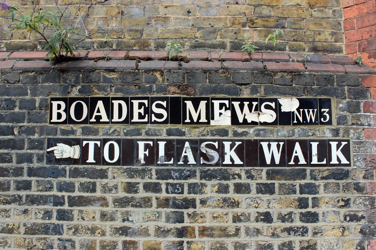 A tiled street sign set in a brick wall in Hampstead reading Boades Mews NW3, with a pointing hand and the words To Flask Walk