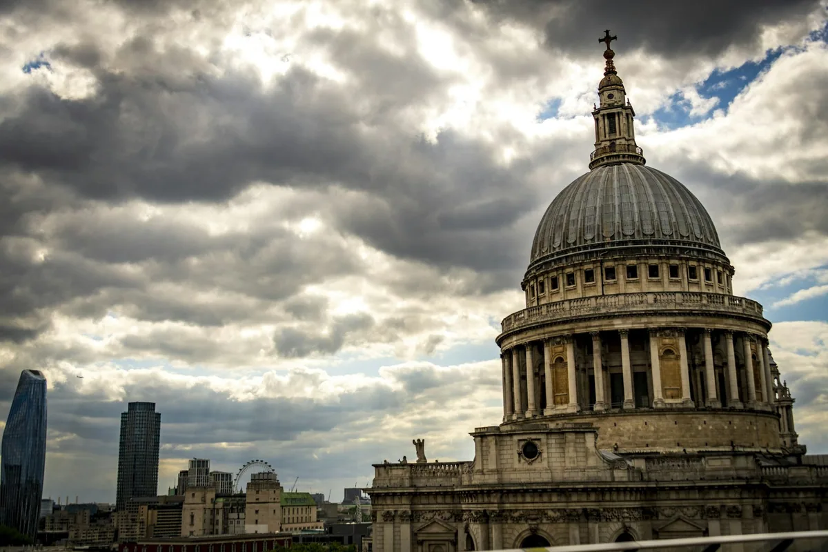 The dome of St Paul's Cathedral under a dramatic cloudy sky, with City towers in the distance