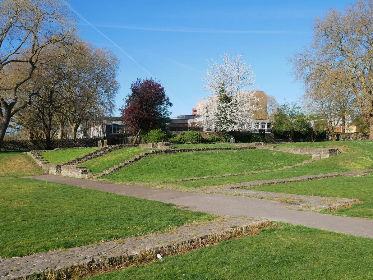 The low stone wall foundations of Barking Abbey laid out in mown grass, paths crossing between them under bare and blossoming trees