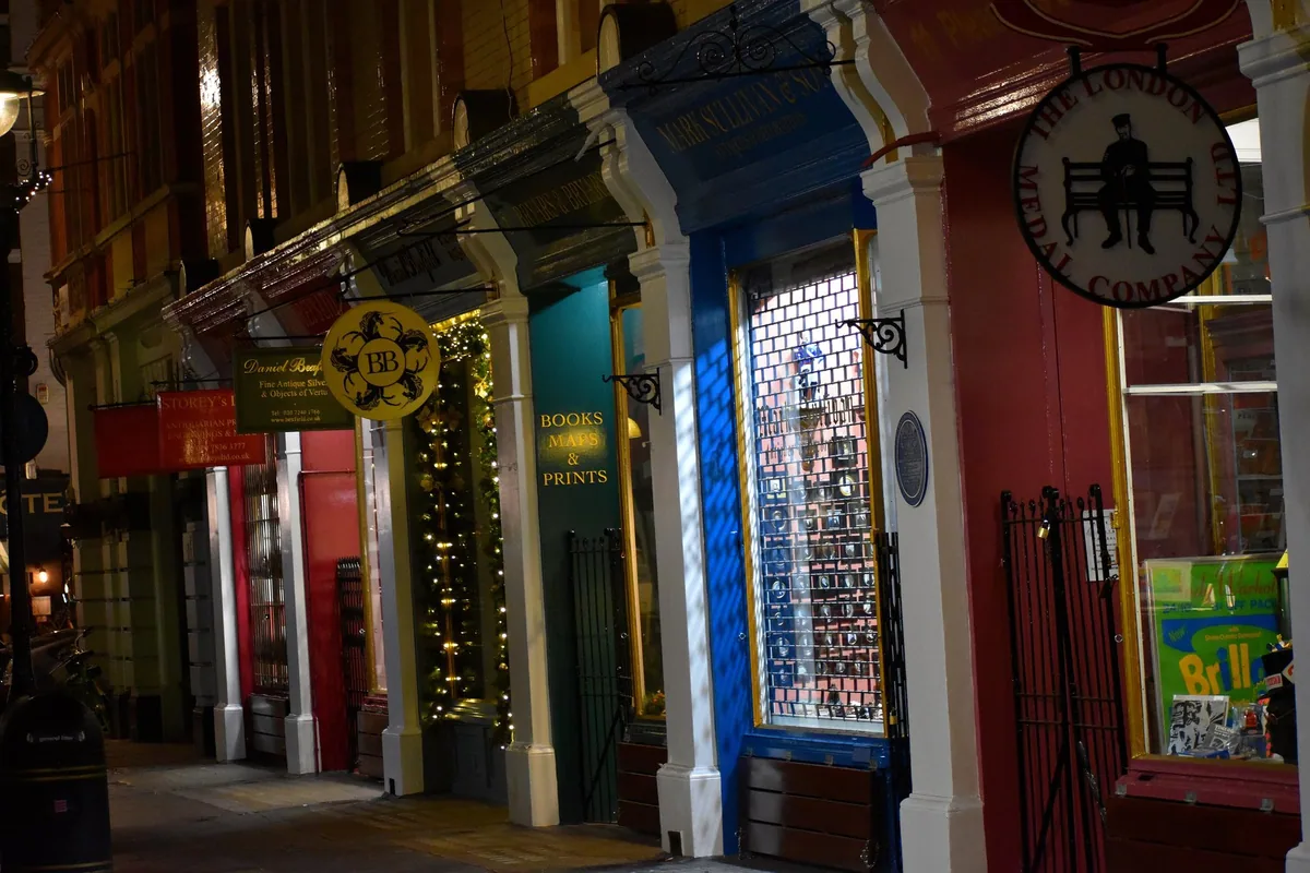 A lit row of antiquarian shopfronts at night, painted green, blue, red and gold, with gilt lettering, a hanging medal-dealer's sign and a window reading Books, Maps and Prints