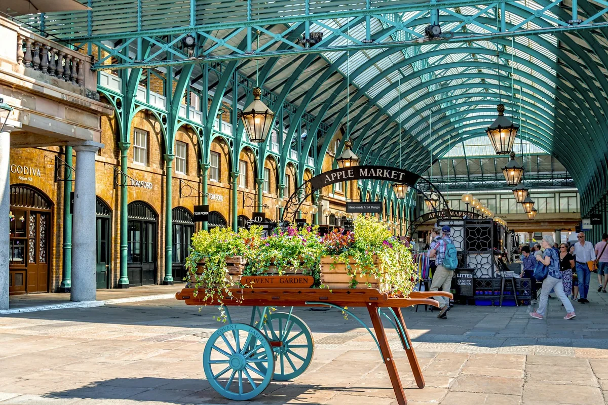 A wooden flower cart under the arched glass roof of Covent Garden Market with shoppers around it