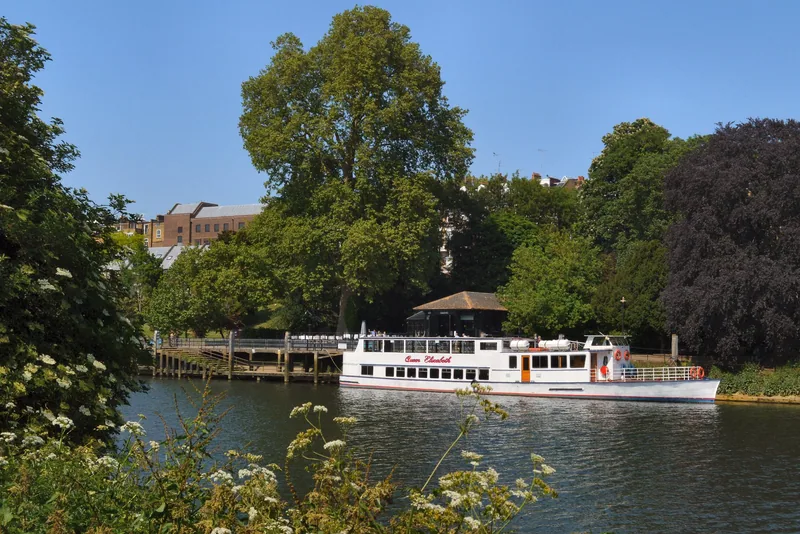 A white pleasure boat moored at the wooded Richmond riverside on the Thames, seen from the water