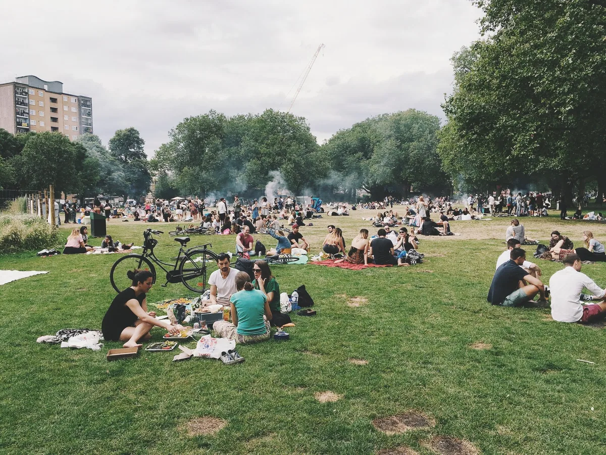 People sitting and picnicking on the grass at London Fields on a sunny afternoon, with tower blocks behind the trees