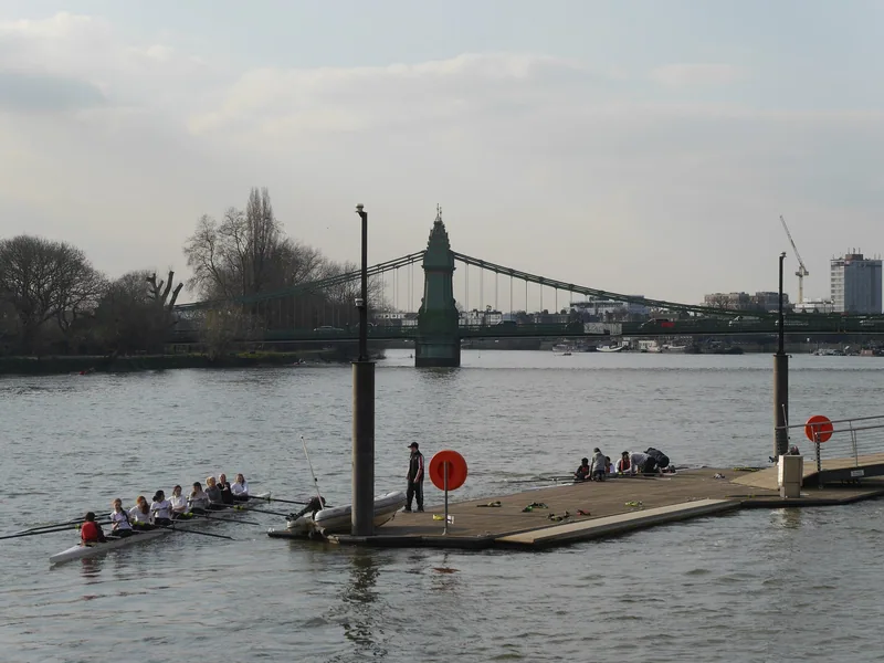 A rowing eight on the Thames passing a landing stage, with Hammersmith Bridge in the distance
