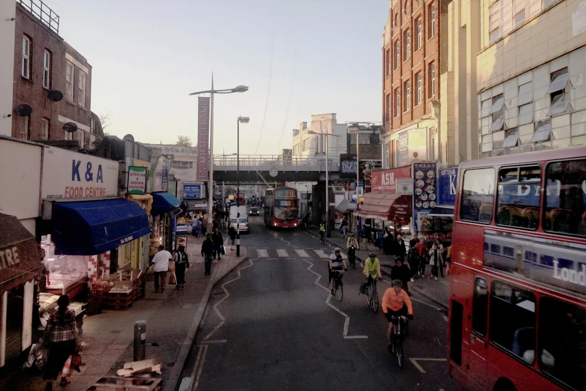 Rye Lane in Peckham in evening light, with shop awnings, cyclists and buses heading for the railway bridge