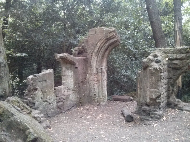 A ruined stone arch with carved mouldings standing among trees in the woodland on Sydenham Hill