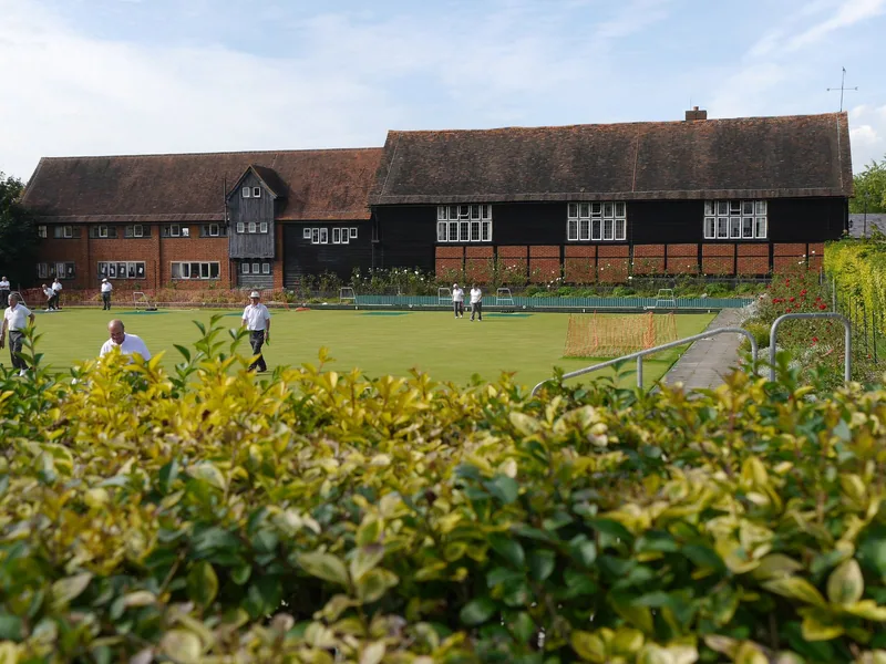 The black weatherboarded timber barns at Manor Farm, Ruislip, seen across a bowling green with a few players on it