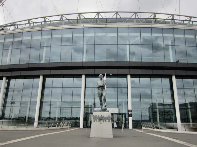 The curved glass frontage of Wembley Stadium with a bronze footballer statue on a plinth outside