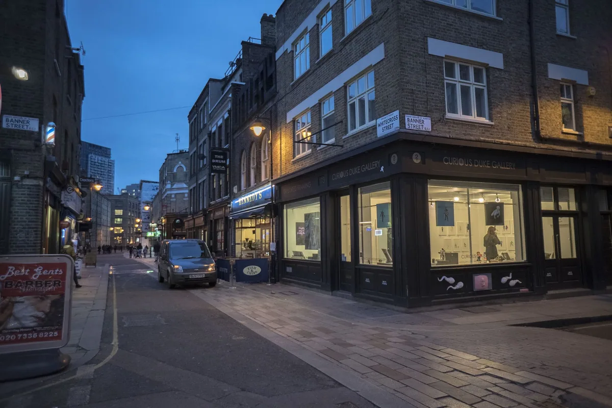 Whitecross Street at dusk after the stalls have gone: lit ground-floor windows along a narrow corner and an empty carriageway