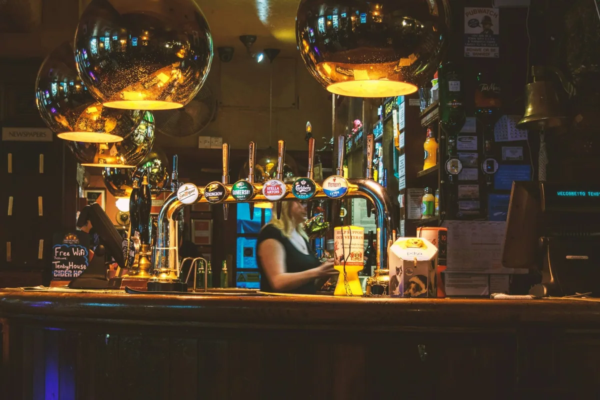 A wooden pub bar lit by brass lamps, with hand pumps along the counter and a bartender working behind it