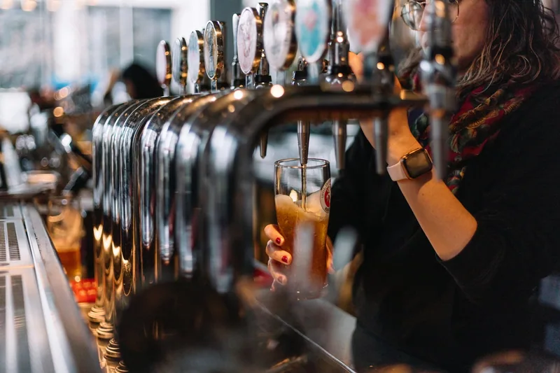 A bartender drawing a pint from a long row of chrome keg taps, the glass half full and held at the spout, decorated tap badges above