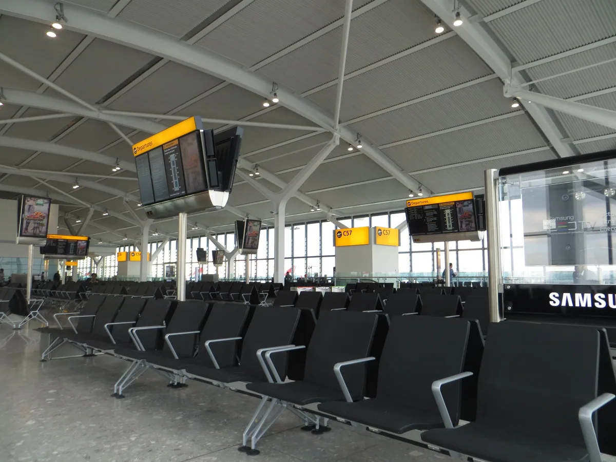 Empty rows of black seats at a Heathrow departure gate, with information screens hanging from the ribbed terminal roof