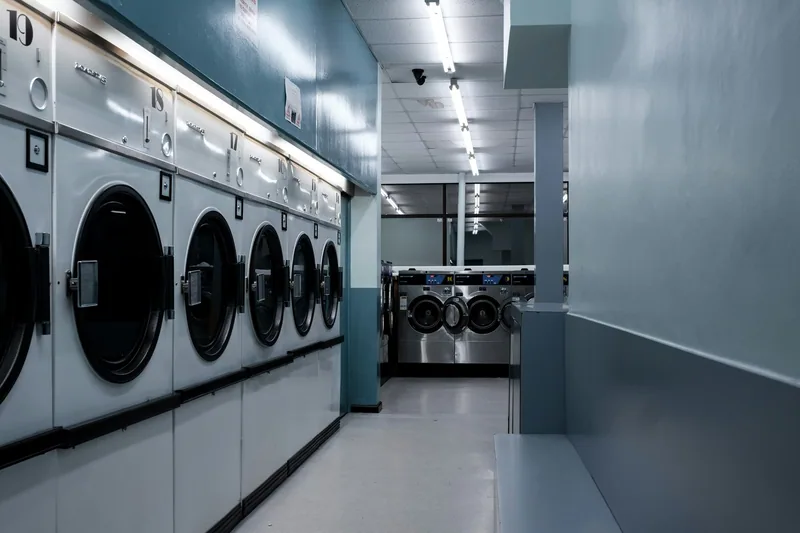 Rows of washing machines and tumble dryers along the wall of an empty launderette under fluorescent light
