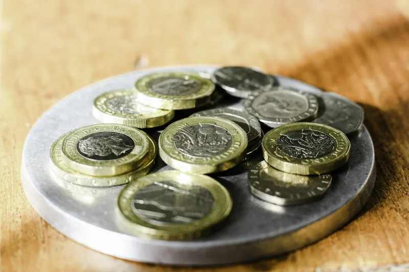 Twelve-sided one pound coins and twenty pence pieces piled on a small metal tray on a wooden counter