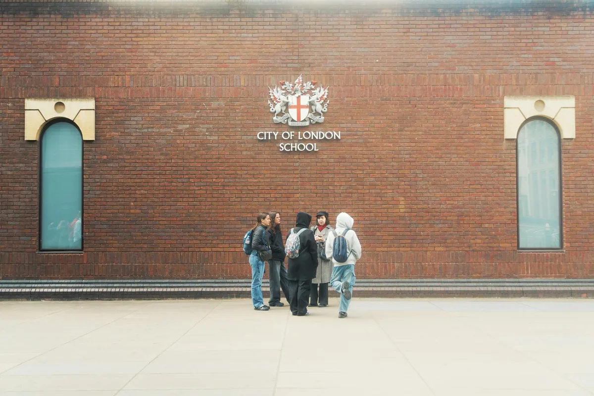 A group of students standing beside the brick wall and carved crest of City of London School