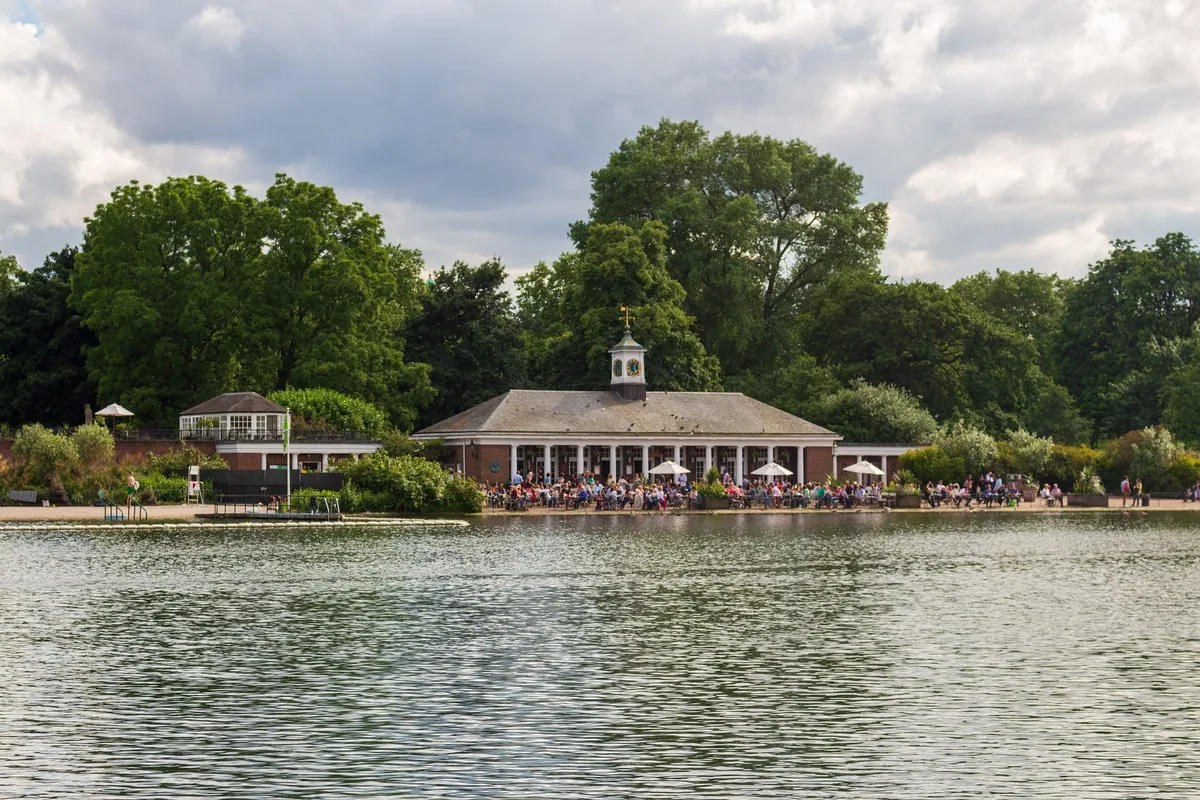 The Serpentine Lido pavilion in Hyde Park seen across the water on a summer day, its terrace busy with people