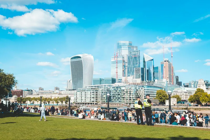 Crowds on a riverside lawn beside the Thames in sunshine, with the City of London skyline rising behind