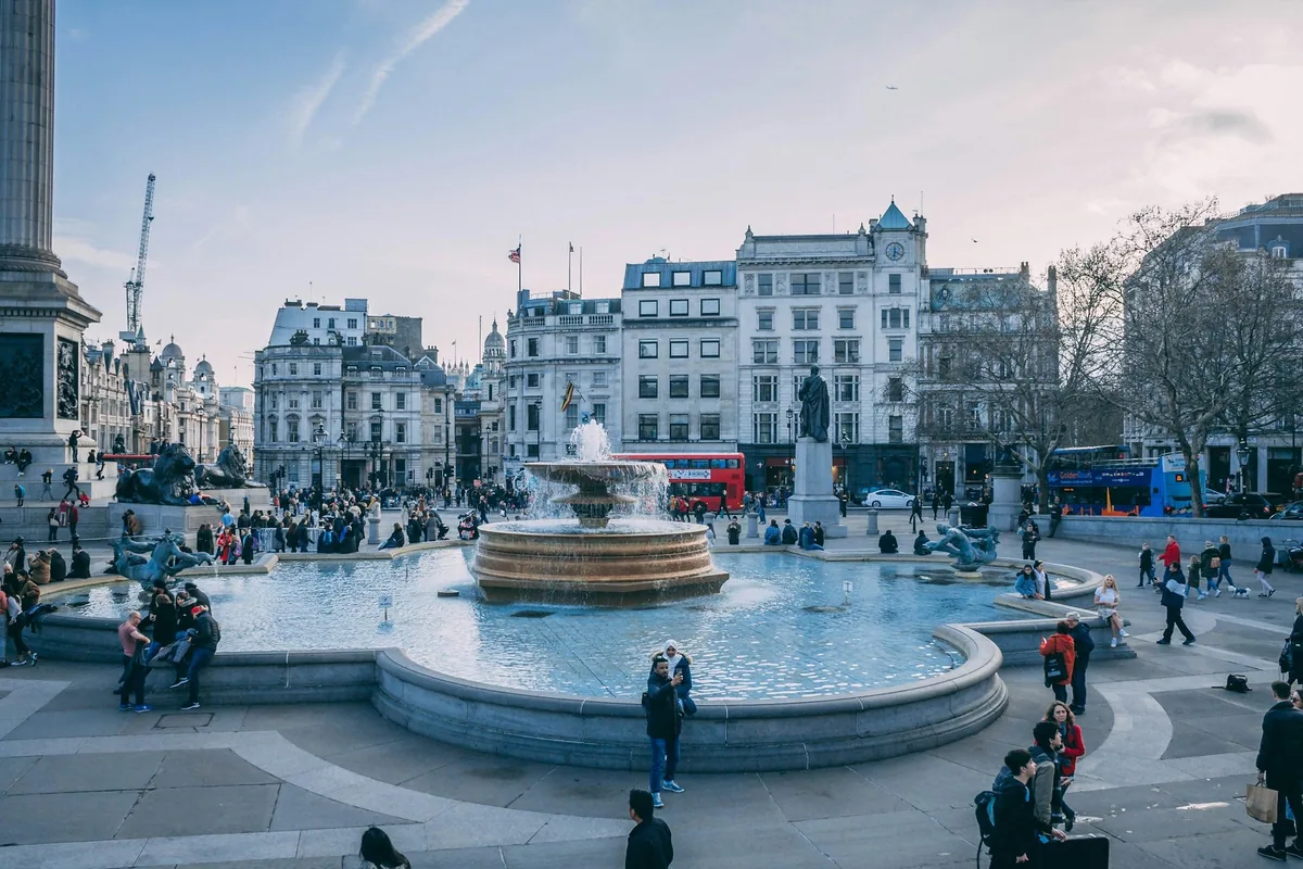 Trafalgar Square on a cold clear day, with the fountain running, bare trees and a red bus passing behind
