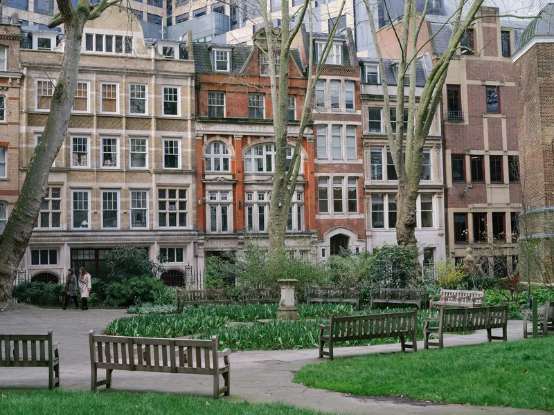 Empty wooden benches and flower beds in a quiet City of London garden, Victorian brick and stone buildings rising behind the bare trees