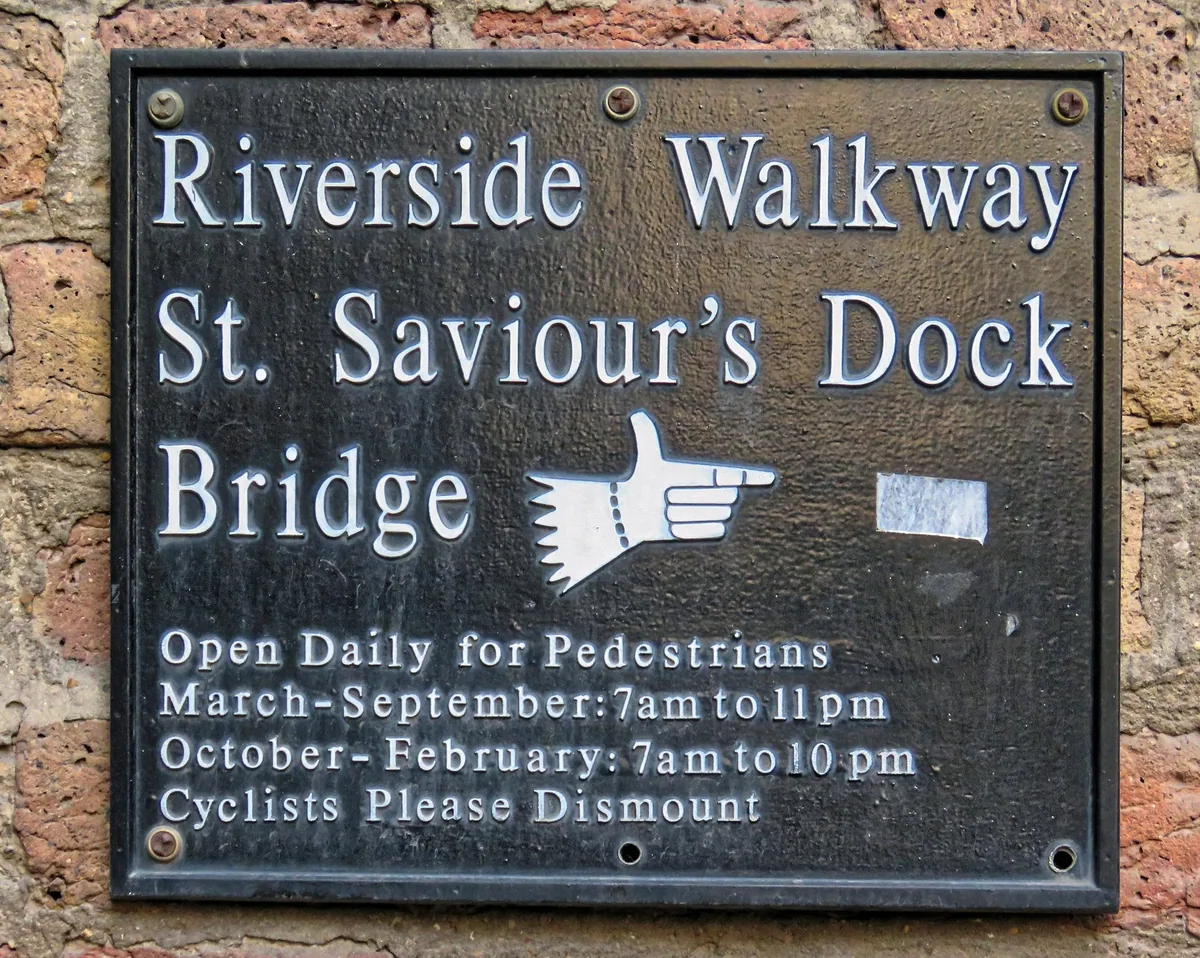 Cast metal sign on a brick wall giving the Riverside Walkway bridge's seasonal opening times for pedestrians