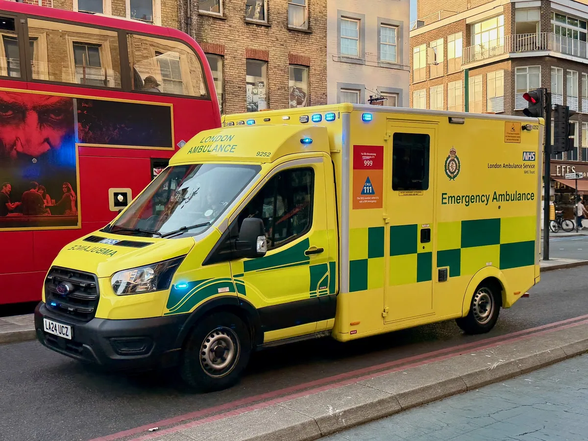 A yellow and green London Ambulance Service emergency ambulance on a city street with a red bus behind