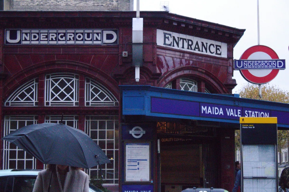 The ox-blood tiled front of Maida Vale Underground station on a wet day, roundel and station canopy above a raised black umbrella