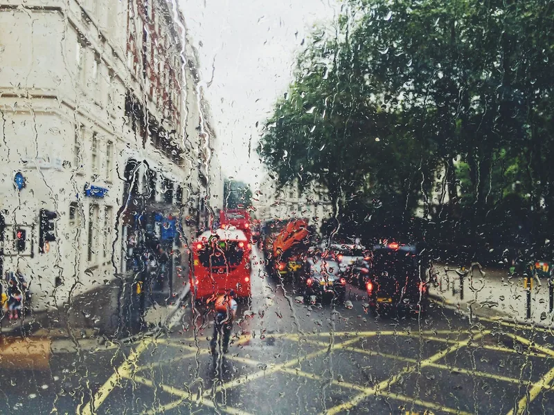 A London street of stucco terraces and a red bus seen through a window streaming with rain