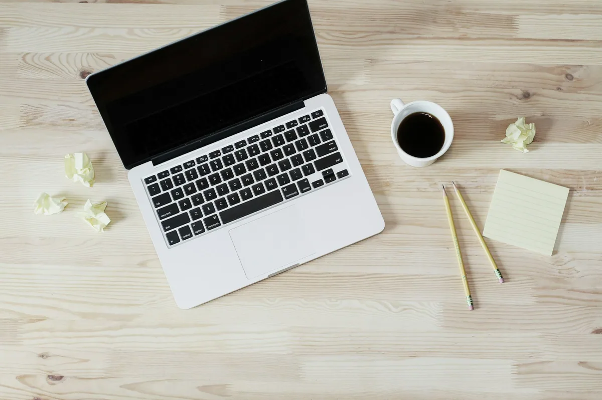 A closed-screen laptop on a light wooden desk beside a cup of black coffee, pencils and sticky notes