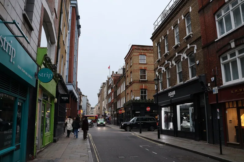 Berwick Street shopfronts with a black cab at the kerb and a red bus at the junction beyond