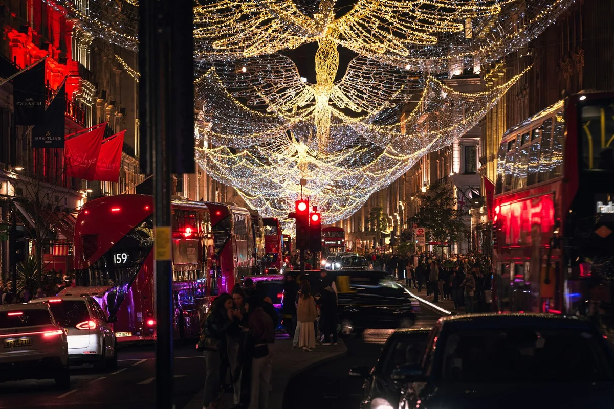 Illuminated angel lights hanging above a London shopping street at night, with red buses and black cabs below