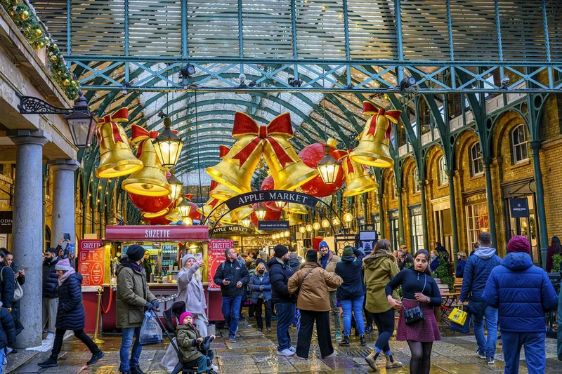 Giant gold bells and red bows hanging under the glass roof of Covent Garden's Apple Market, with crowds below