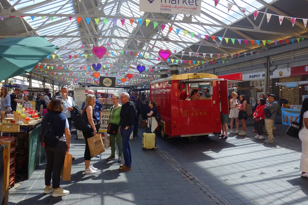 Shoppers among stalls under the glass roof and bunting of Greenwich Market