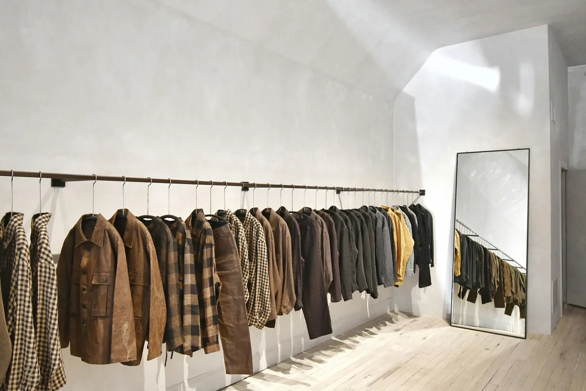 A long rail of jackets and shirts along a bare white wall beside a leaning floor mirror