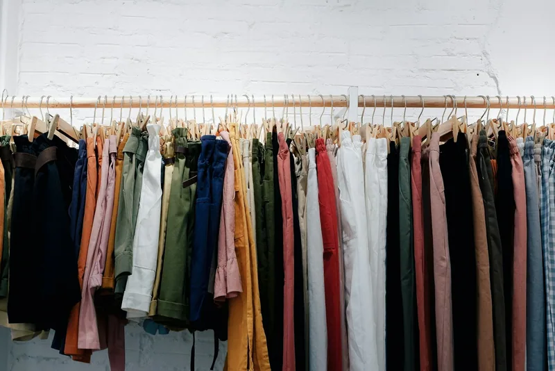 Trousers in many colours packed tightly on a wooden rail against a whitewashed brick wall
