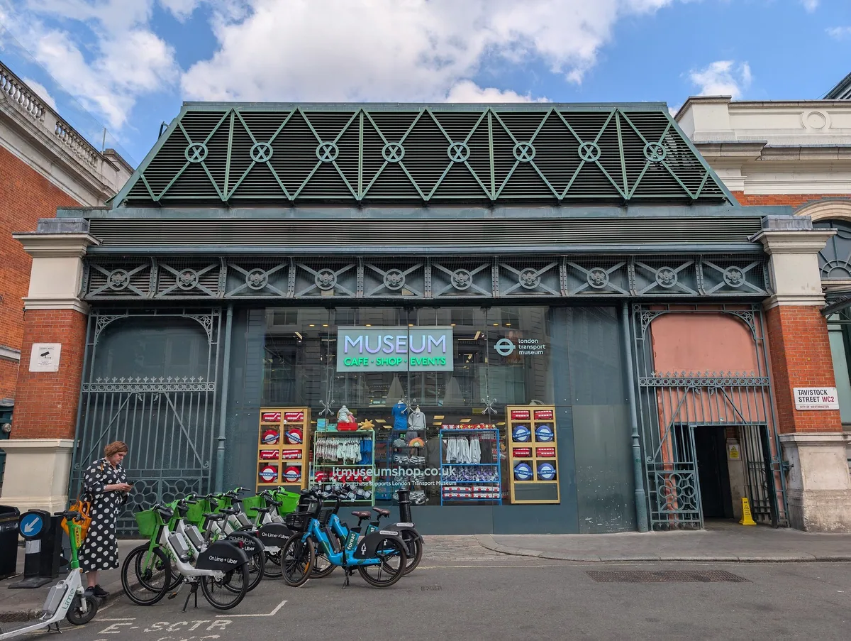 The street-facing glass shopfront of the London Transport Museum shop under a green iron market roof, posters in the window
