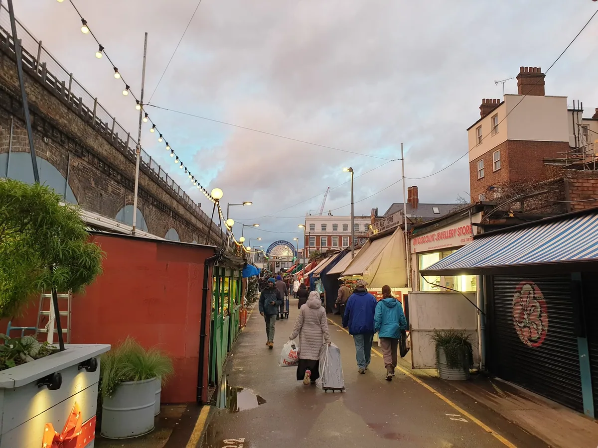 The alley of Shepherd's Bush Market between stall awnings and festoon lights, with the railway viaduct running along the left side