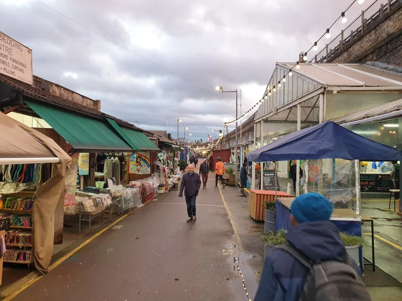 Fabric, clothing and household stalls along Shepherd's Bush Market, with green awnings and shoppers on the narrow lane