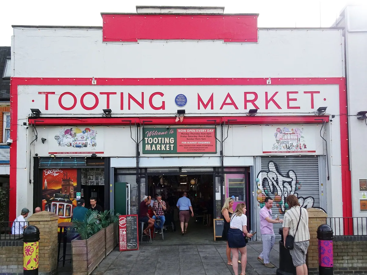 The red and white entrance front of Tooting Market on Totterdown Street, with shoppers outside