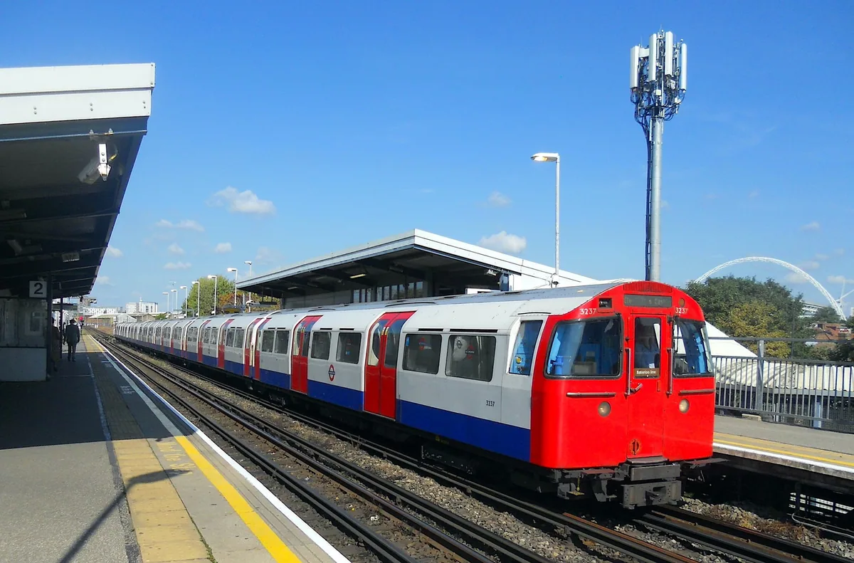 A red and white 1972 stock Bakerloo line train arriving at an open-air platform under a blue sky