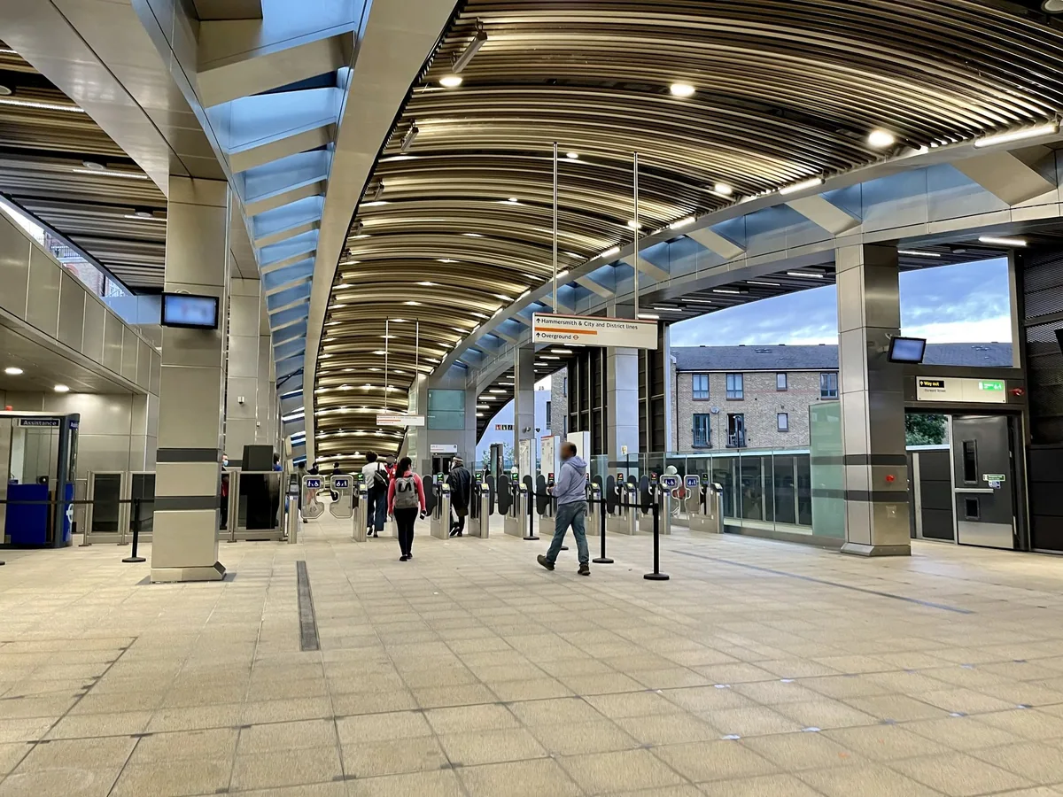 Passengers crossing the wide ticket hall at Whitechapel station towards the row of gates, under a curved timber ceiling