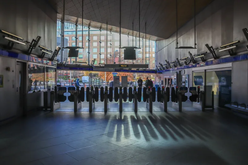 A row of ticket gates inside Wood Lane station, sunlight falling through the glazed wall behind them