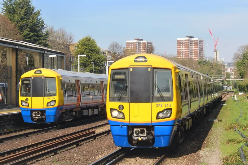 Two London Overground Class 378 trains standing at Kensington (Olympia) station, with tower blocks rising behind the tracks