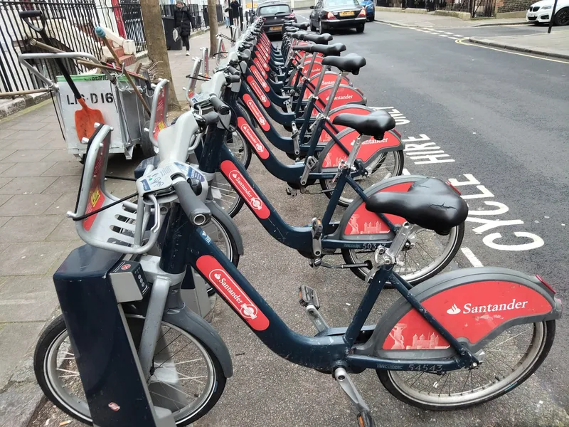 A long row of red Santander hire bikes at a kerbside dock marked Cycle Hire
